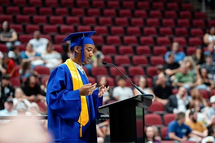 A graduate wearing a blue cap and gown stands at a podium, delivering a speech during a graduation ceremony as the audience looks on.