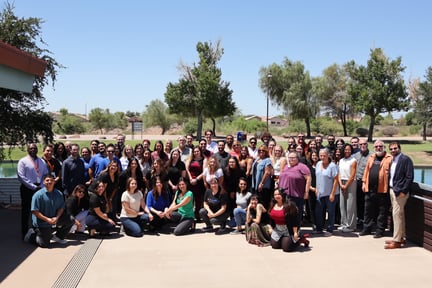 A large group of staff is standing and sitting together outside near a pond and trees