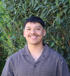 A headshot of Miguel Acosta in front of a bush